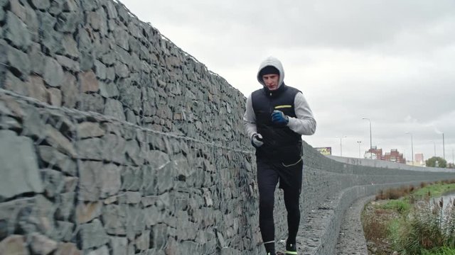 Dolly shot with slow motion of young man running near river alongside stone wall on cold day