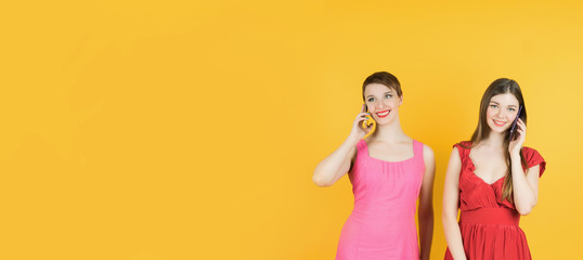 Technology, friendship and leirure concept - two smiling young women talking with smartphones. Isolated studio, yellow background, female model. Banner.