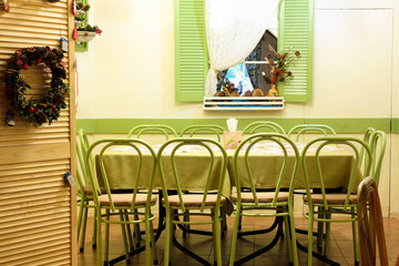Light green chairs in a row, a table, a window with green shutters, white walls and dim light bulbs in cafe bar. Interior of the cafe