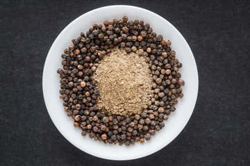 Black pepper grains with ground pepper in the white dish on the black background. Composition of seasoning. Healthy eating and lifestyle.