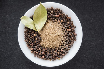 Black pepper grains with ground pepper in the white dish on the black background. Composition of seasoning. Healthy eating and lifestyle.