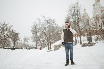 Outdoor winter portrait for young handsome man. Beautiful teenager in his jacket and vest posing on a city street, background of fir trees.