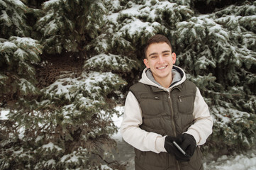 Outdoor winter portrait for young handsome man. Beautiful teenager in his jacket and vest posing on a city street, background of fir trees.