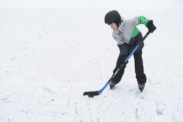 Child boy playing ice hockey at the skating rink outside in winter.