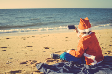 Fototapeta na wymiar Child boy Santa Claus using mobile phone on ocean beach outdoors background, back view photo.