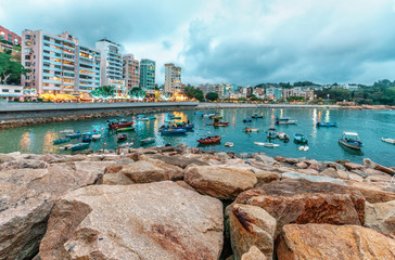 Fototapeta premium Rocky sea shore of Stanley Bay on Hong Kong Island in Hong Kong. Beautiful scenic landscape with water, mountains, rocks, buildings, fishing boats and cloudy sky