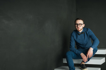 studio portrait of a young man on a dark wall background in a blue shirt and jeans. hipster man, dressed stylishly, with a beard and glasses, sitting on white stairs