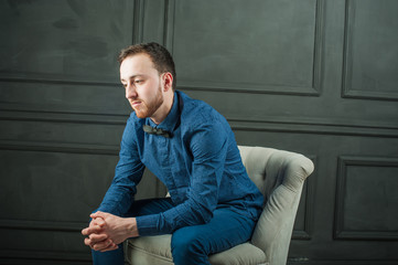 studio portrait of a young man on a dark wall background in a blue shirt and jeans. hipster man, dressed stylishly, with a beard, sitting in a light easy chair