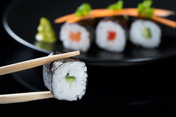 Sticks holding roll with cucumber on the background of fresh rolls with salmon, cucumber and pepper on a plate, served with carrot slices and parsley, black background