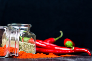 Chili powder, dry basil and chili peppers on black table. Paprika, oregano and red chili peppers in glass jars on dark wood background