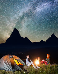Night tent camping. Happy couple tourists sitting by campfire under incredibly beautiful starry sky and Milky way. In the background silhouette of the high mountains and luminous village in the valley