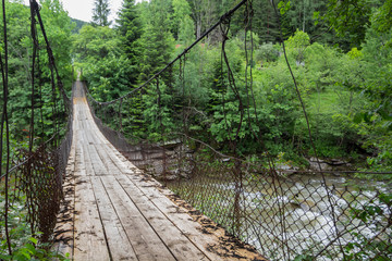 Old suspension bridge over the river in the forest. - obrazy, fototapety, plakaty