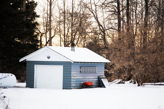 A red wheel barrow beside a painted weathered one car garage with attached ladder in yard sire in winter