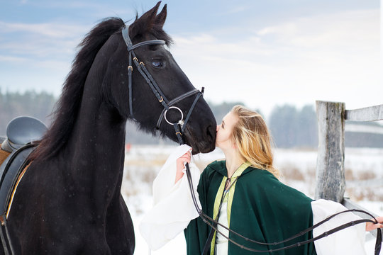 Young girl in white dress with black thoroughbred horse on winter field. Historical equine background