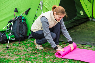 Cheerful woman rolling camping mat , during the camping holidays. In the background tourist tent...