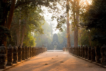 Angkor Wat Cambodia temple statue avenue woman sweeps the floor at sunrise golden light ambience with trees
