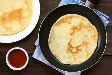 Freshly made crepe in frying pan with a stack of crepes and strawberry jam on the side, photographed overhead with natural light