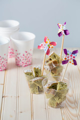 Japanese matcha green tea cake pieces served in individual glasses. Dessert is topped with colorful paper pinwheels. Light wood background. 