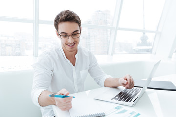 Young cheerful man using laptop while writing notes