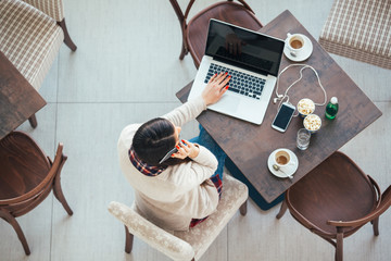 Freelance young woman sitting in the cafeteria with laptop and using mobile phone