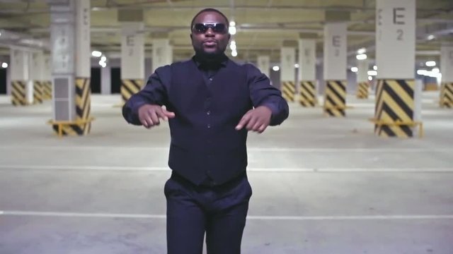Dolly shot of African man in suit and sunglasses walking through parking lot and rapping 