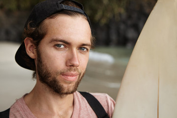 Close up portrait of young handsome winner of surf contest, looking at camera with sly satisfied smile, holding his lucky board, isolated on blurred background of sea beach. Active summer recreation