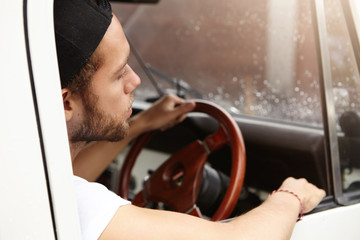 Close up shot of attractive young Caucasian hipster wearing t-shirt and baseball cap backwards, sitting inside his white safari vehicle with hand on wheel, driving along rural road. View from back
