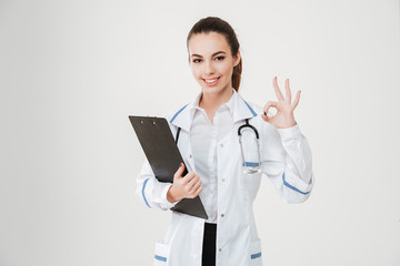 Cheerful beautiful young woman doctor with clipboard showing ok sign