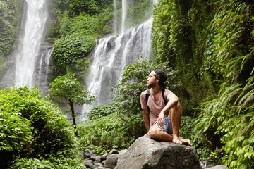 Fototapeta na wymiar Nature, tourism and people. Young barefooted tourist wearing jeans short and backpack sitting on big rock deep inside green rainforest, looking peaceful and relaxed with waterfall on background