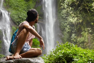 People, lifestyle and adventure concept. Stylish young man wearing cap backwards sitting on big rock, contemplating wonderful waterfall in green rainforest. Barefooted hiker trekking in jungle