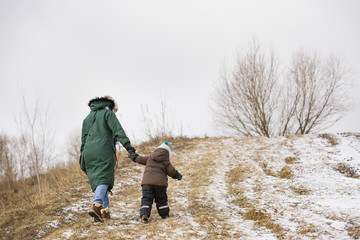 Small toddler boy and his mother walking the mountain trail on a winter day. Woman with little kid walking up the hill on a windy day. Boy following his mom on the valley trail.
