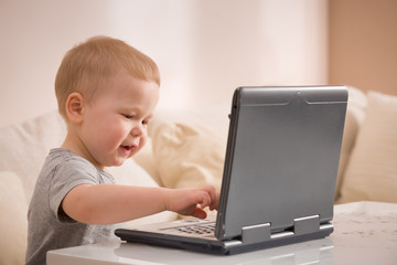 Cute little toddler boy sitting on the sofa with his laptop and looking at the window. Learning with computer. Early development. Child and computer.