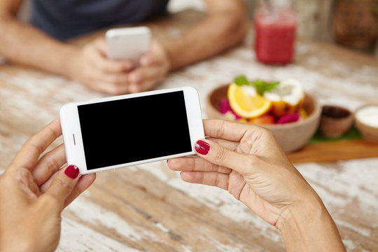 Young woman with beautiful red nails holding mobile phone horizontally with both hands while watching movie or viewing pictures online via social media websites during breakfast at cafeteria