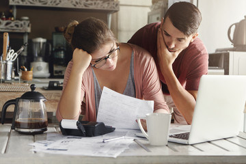 Young stressed Caucasian couple facing financials troubles, sitting at kitchen table with papers,...
