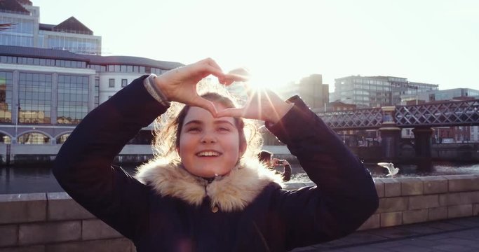 Beautiful girl smiling and making heart with her hands over river background. The sun shines girl's heart and hair. Winter holidays. Slow Motion 4k.