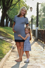 Pregnant mother and her little happy daughter stand in sunny par