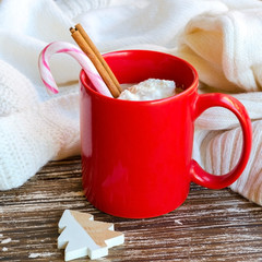 Red mug of hot chocolate with cinnamon stick  and candy cane, white cozy knitted sweater on the back. Delicious cold weather beverage for winter times concept.