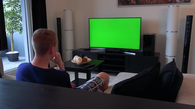 A man watches a TV with a green screen in a cozy living room, eventually looks away