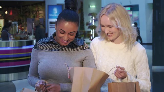 Two pretty women looking into the shopping bag at the mall. Close up of beautiful african american and caucasian girls having fun at the shopping. Attractive blond lady showing her purchases to her