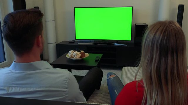 A man and a woman (the backs of their heads to the camera) watch a TV with a green screen in a cozy living room
