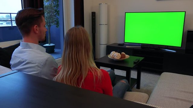A young, attractive couple watches a TV with a green screen in a cozy living room, then turns to the camera and smiles