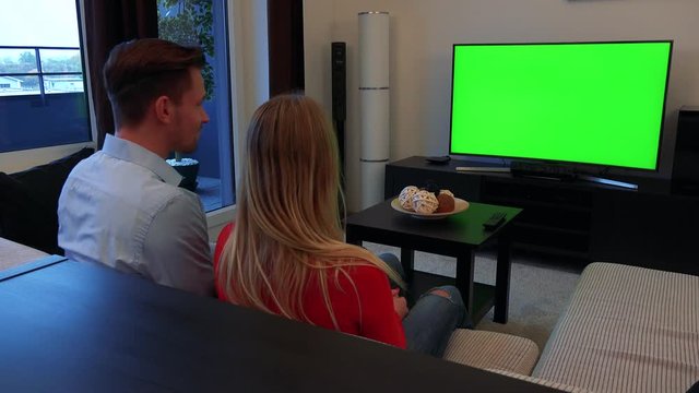 A young, attractive couple watches a TV with a green screen in a cozy living room, turns to the camera and shows thumbs up with a smile