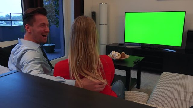 A young, attractive couple watches a TV with a green screen in a cozy living room and celebrates