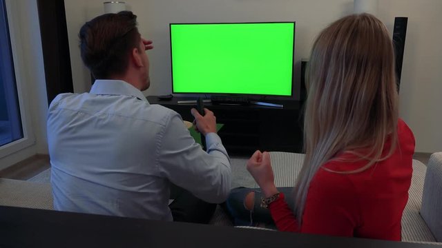 A couple (the backs of their heads to the camera) cheers on and watches a TV with a green screen in a cozy living room, then celebrates