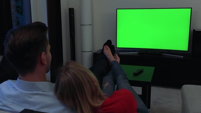 A couple (the backs of their heads to the camera) watches a TV with a green screen in a cozy living room, feet on a table