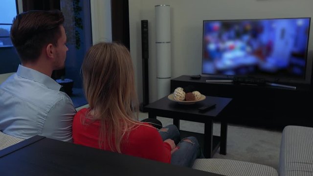 A young, attractive couple watches a TV in a cozy living room, then turns to the camera and smiles