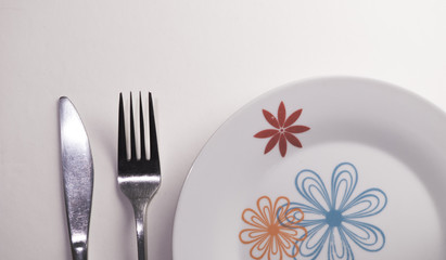 Knife and fork laying next to a plate with flowers painted on it on a white background