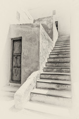 A sepia view of a stairway leading to the upper story of a traditional Arabian house with an old wooden storeroom door.