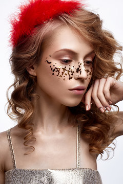 Beautiful girl in a gold dress with a gentle make-up. Model with red feathers on her head and curls. Holiday photo. The beauty of the face. The photo was taken in a studio.