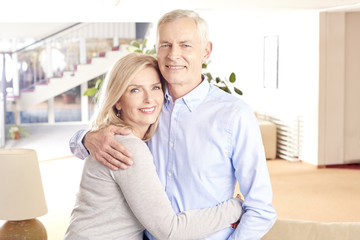 Lovely senior couple. Shot of a happy senior couple standing together. Elderly man embracing her wife from behind while at home in living room.